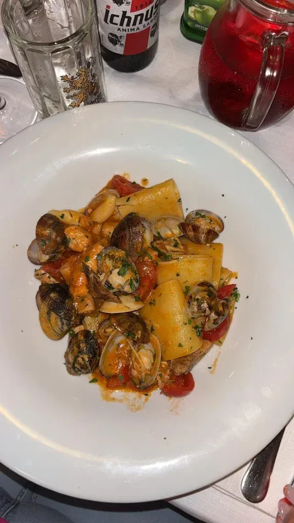 Un piatto di pasta con vongole e pomodori, accompagnato da una bottiglia di birra e una brocca di liquido rosso su un tavolo.