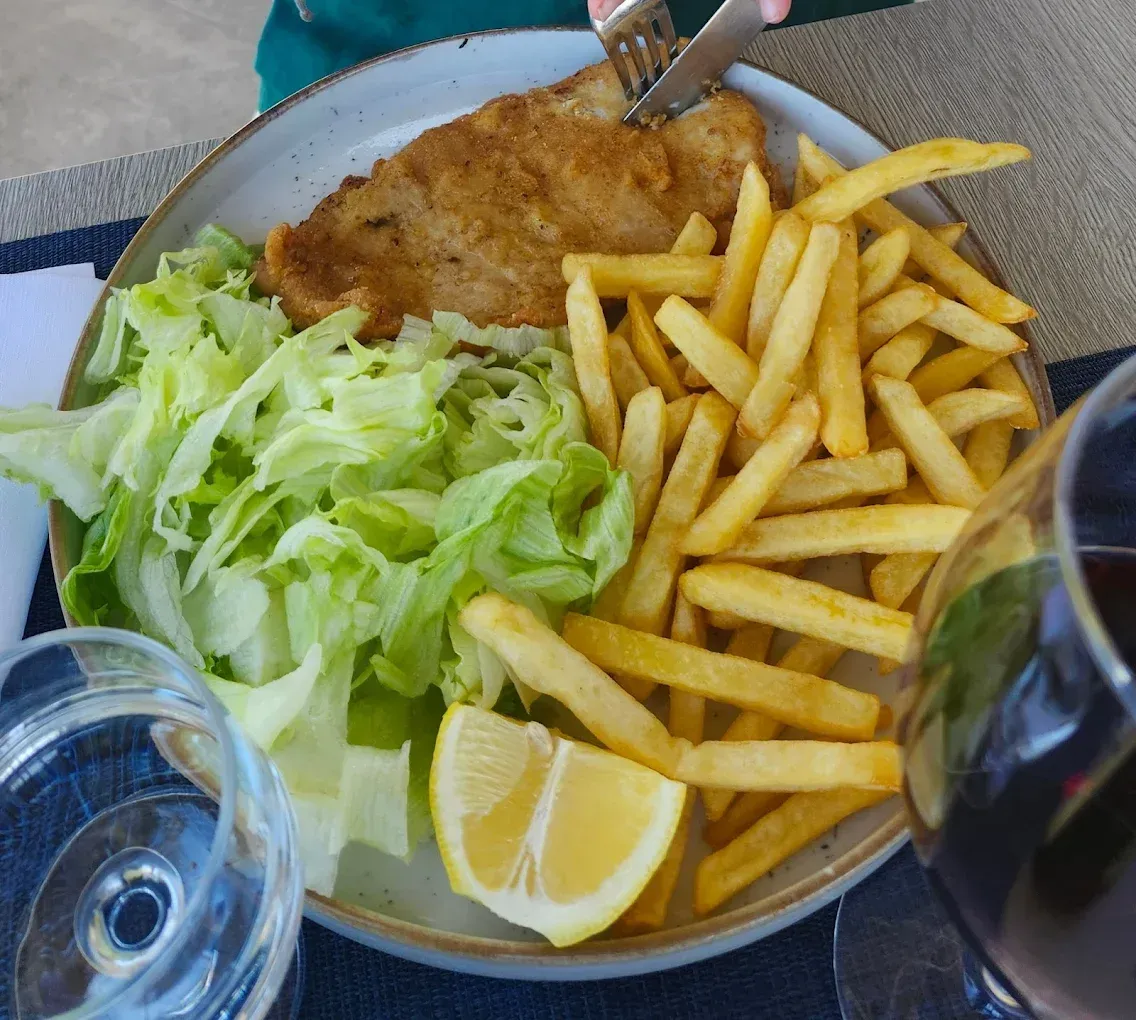 Un piatto con cotoletta impanata, patatine fritte, un'insalata di contorno e uno spicchio di limone, con le mani che tagliano la carne.