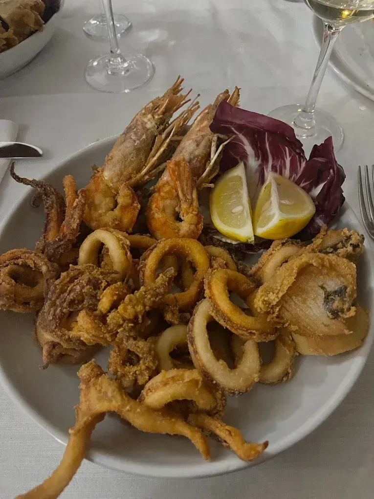 Un piatto di frutti di mare fritti, tra cui anelli di gamberi e calamari, servito con spicchi di limone e radicchio su un tavolo bianco.