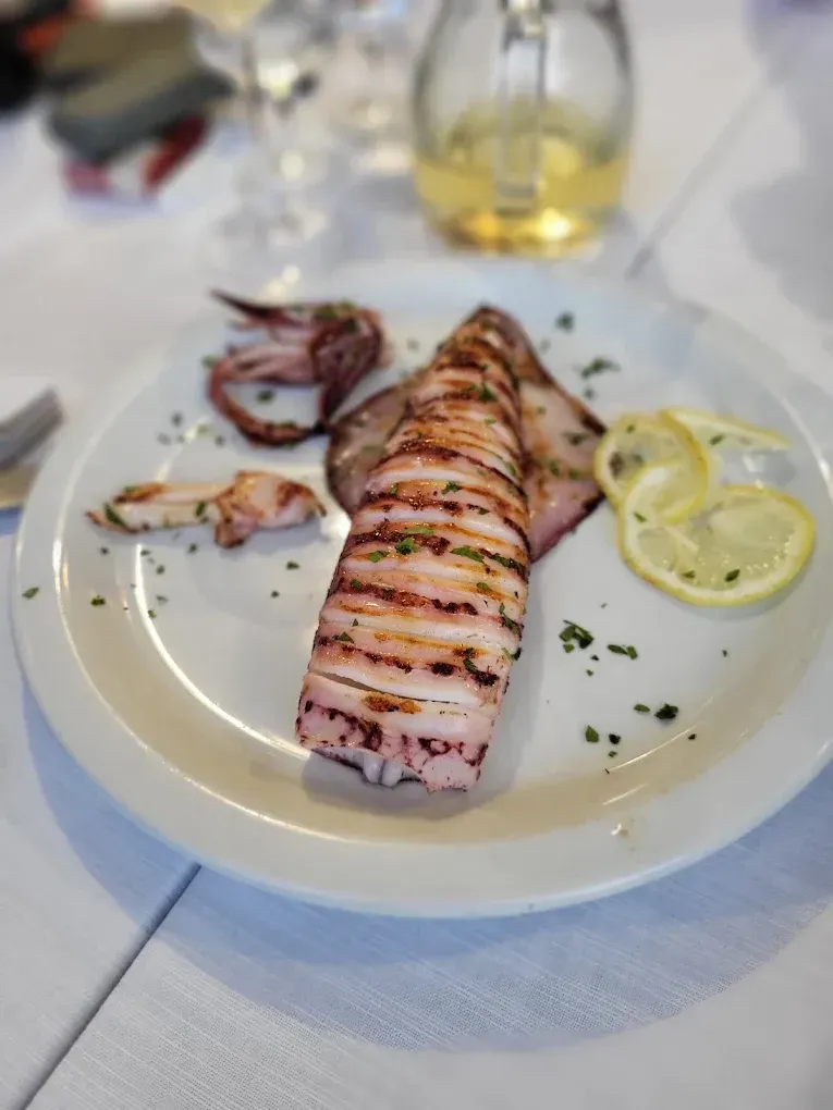 Un piatto di calamari grigliati e affettati, servito su un piatto bianco con fette di limone ed erbe aromatiche, su un tavolo di ristorante.