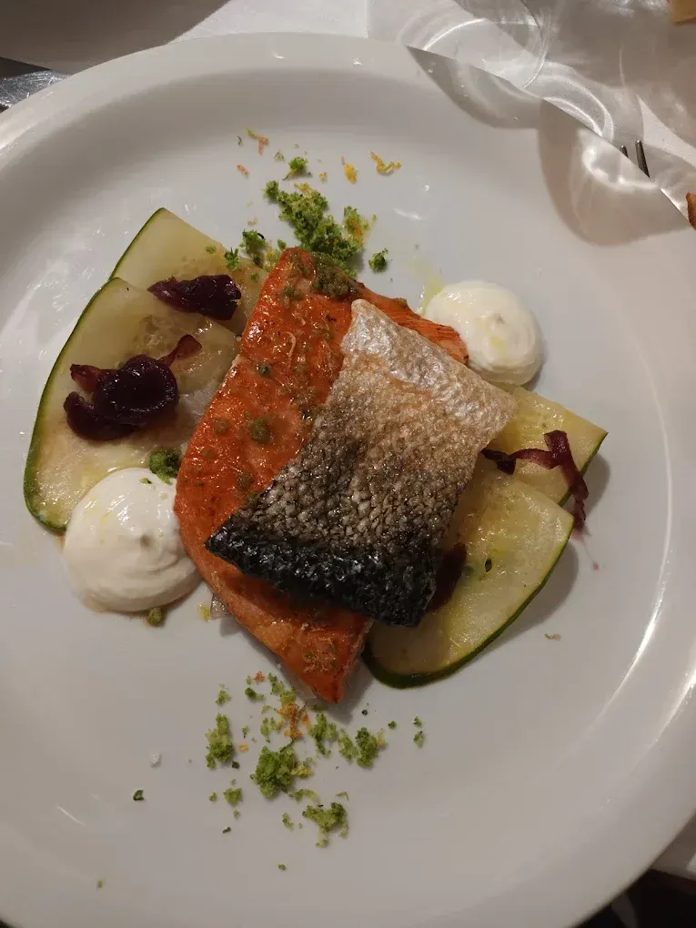 Un piatto composto da un filetto di salmone dalla pelle croccante, zucchine a fette, una guarnizione scura e due cucchiaiate di panna bianca.