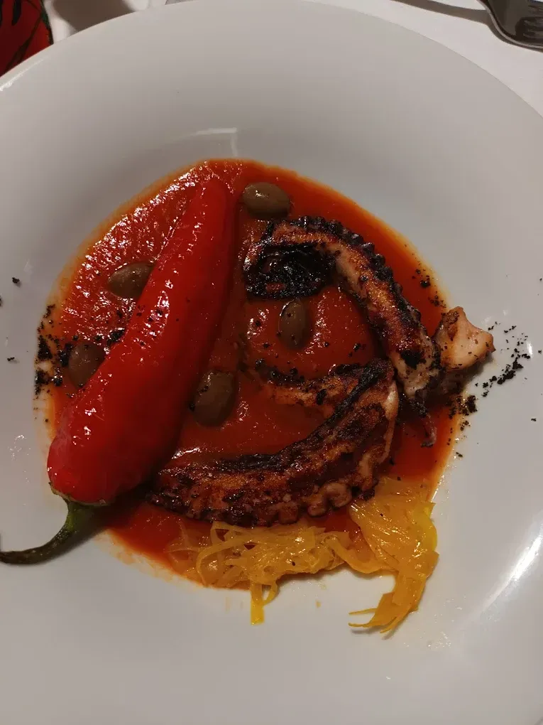 Tentacolo di polpo grigliato servito in salsa di pomodoro con olive, un peperoncino rosso intero e guarnizione di cavolo cappuccio giallo tritato.