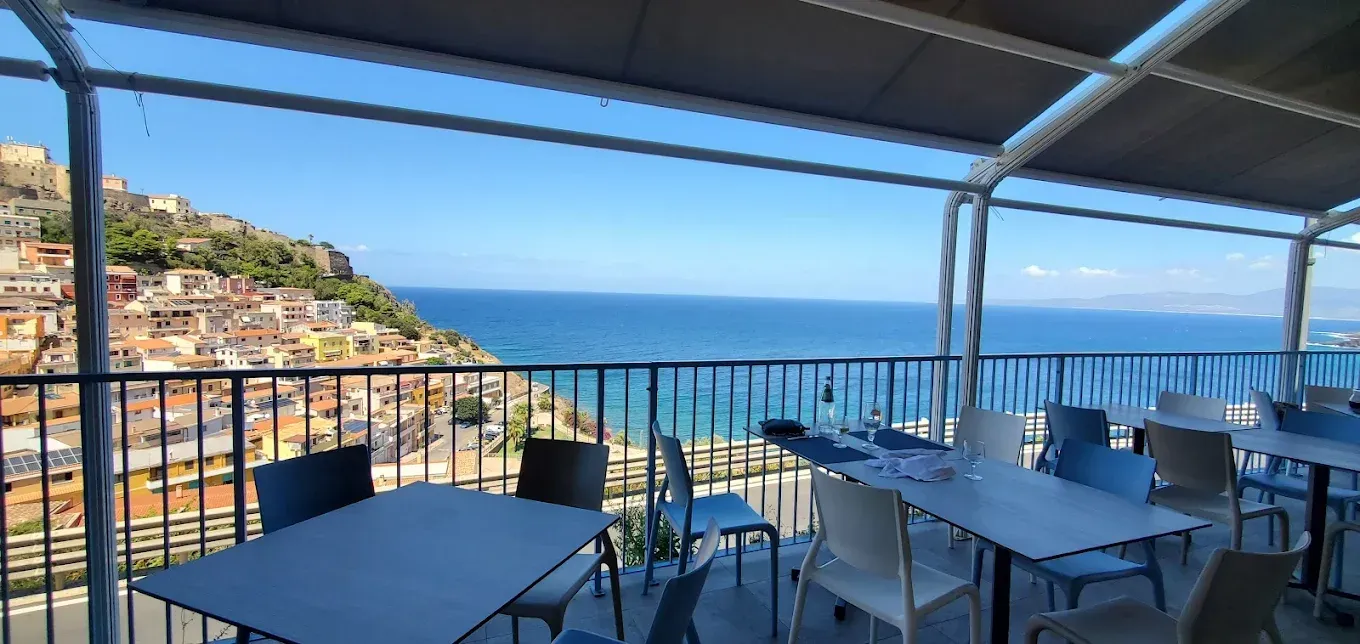 Terrazza esterna del ristorante con tavoli e sedie, affacciata su una cittadina costiera e sul mare, sotto un cielo azzurro e limpido.