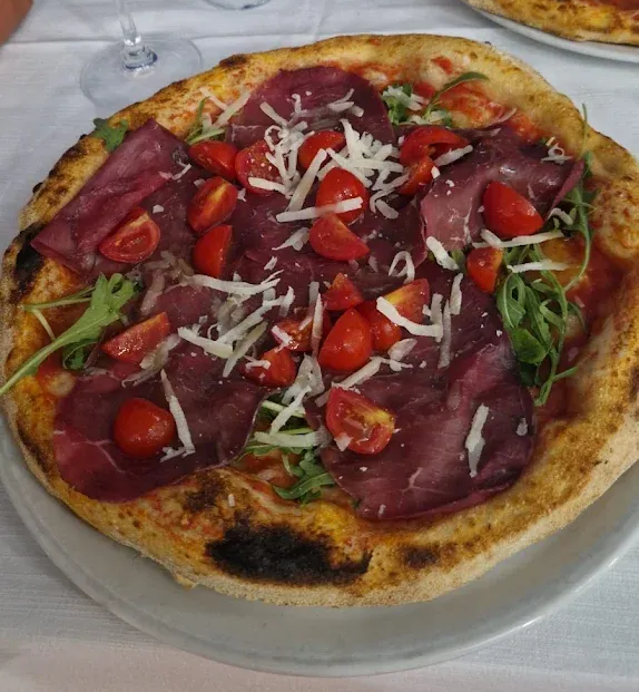 Una pizza bresaola con rucola fresca, pomodorini ciliegino tagliati a metà e scaglie di parmigiano, servita su un piatto bianco.