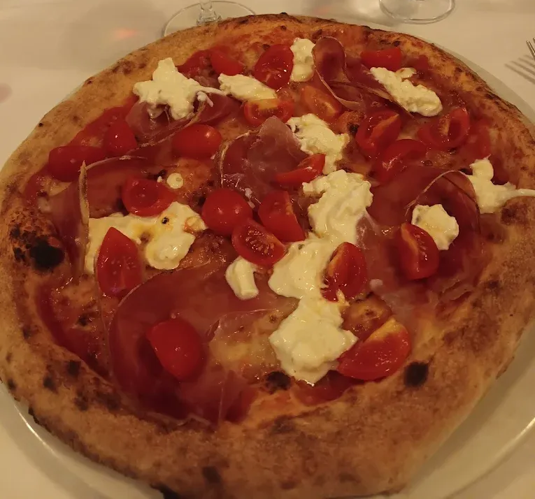 Una pizza a crosta sottile condita con prosciutto crudo, pomodorini freschi e abbondanti cucchiaiate di formaggio bianco, servita su un piatto bianco.