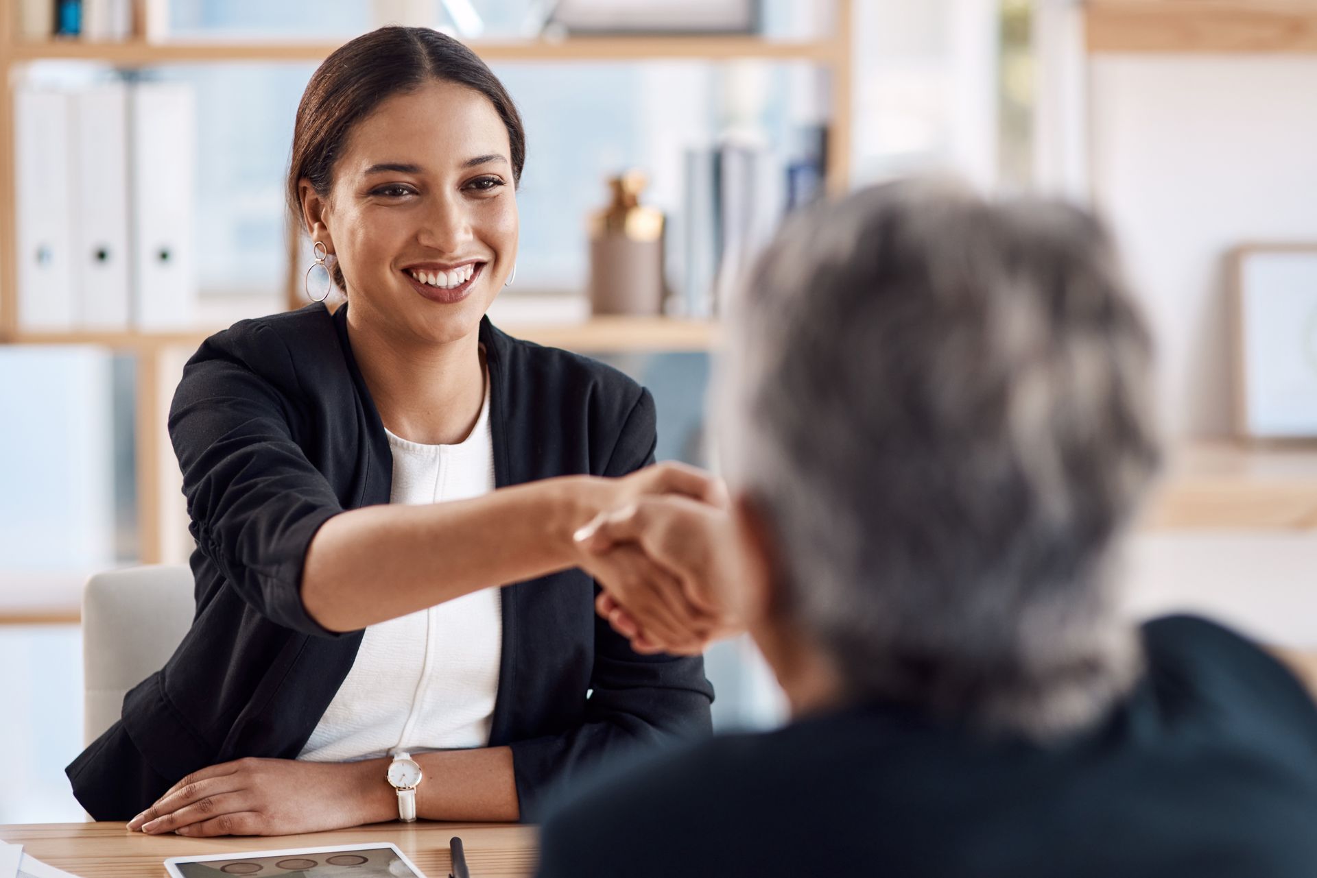 Woman in black blazer shaking hands with another person, smiling. Office setting.