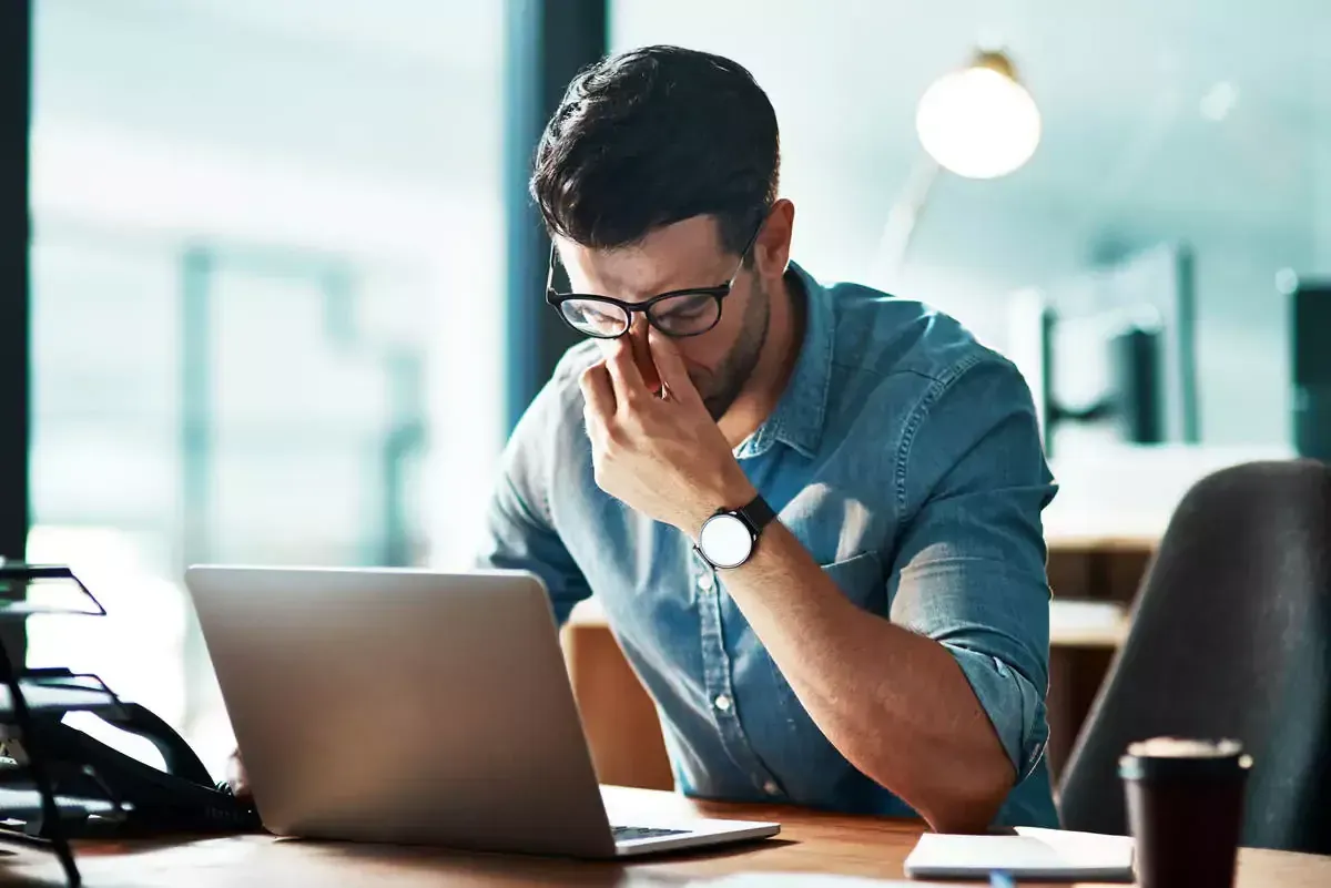 Man wearing glasses, rubbing his eyes while working on a laptop in an office setting.