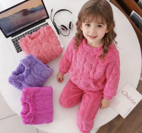 Una niña con un conjunto de punto rosa está junto a un ordenador portátil, unos auriculares y unos jerséis doblados sobre una mesa redonda blanca.