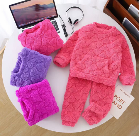 Conjunto infantil rosa de forro polar con capucha y pantalón, además de piezas dobladas a juego de color morado y magenta sobre un escritorio.