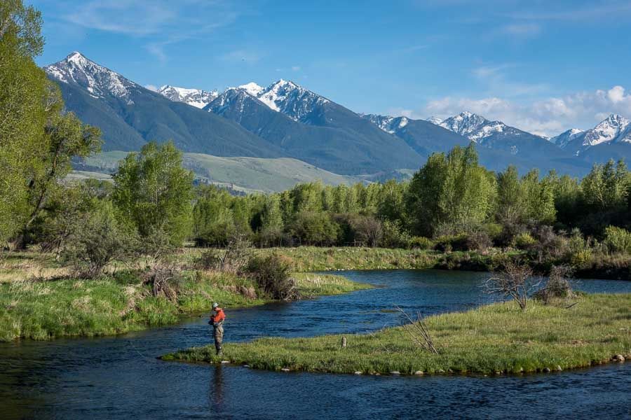 A man is fishing in a river with mountains in the background.