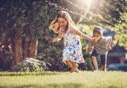 Children playing in the sprinkler on a sunny lawn; girl in floral dress, boy in shorts, laughing.