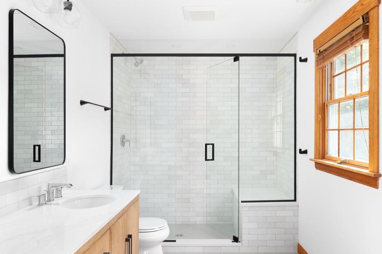White bathroom with a black-framed shower, a vanity, and a window with a wooden frame.