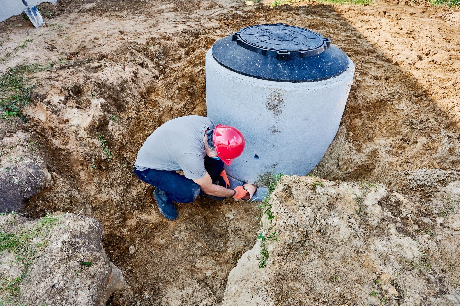 Man in red helmet working on a concrete septic tank in a dirt pit outdoors.