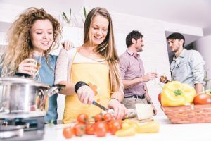 Friends cooking together in a kitchen: woman slicing tomatoes, others chatting, drinking, and smiling.