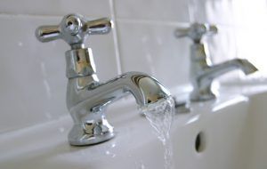 Chrome faucet with water running into a white sink, another faucet visible in background.