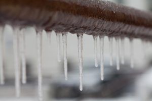 Icicles hanging from a brown pipe, partially blurred background, suggesting a cold outdoor environment.