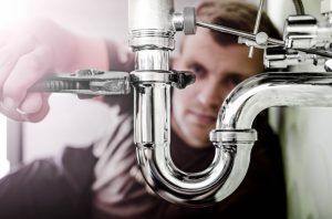 Plumber tightening a shiny metal pipe with a wrench; indoor setting.