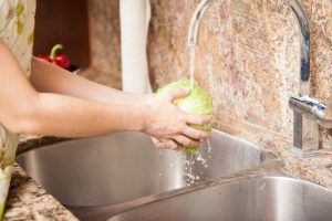 Person washing a green cabbage in a stainless steel kitchen sink under running water.