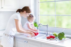 Woman and child wash vegetables in a kitchen sink near a window.