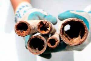 Gloved hands holding sections of corroded, rusted pipes, showing mineral buildup and decreased water flow.