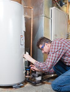 Man in plaid shirt and safety glasses inspects a water heater in a utility room.