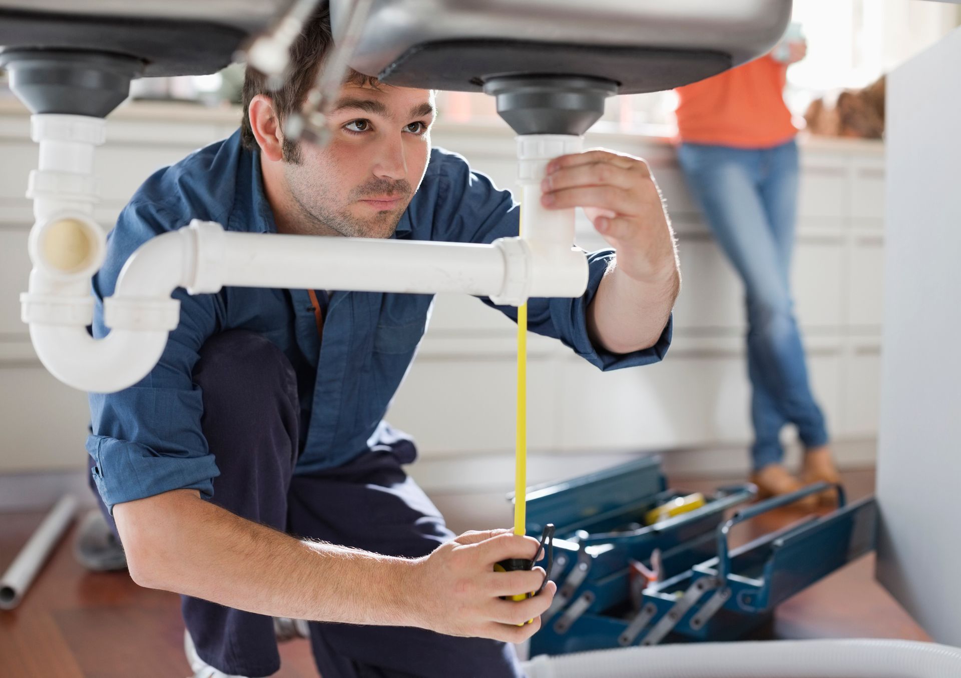 Plumber measures pipe under a sink with a tape measure. Woman in background.