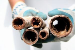 Hands in gloves hold corroded pipes, showing rust and mineral buildup inside.