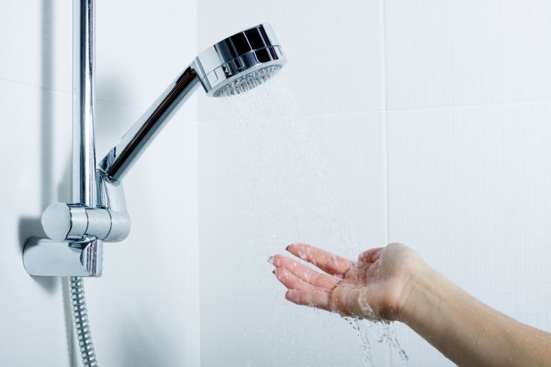 Hand under shower head, testing water temperature against a white tiled wall.