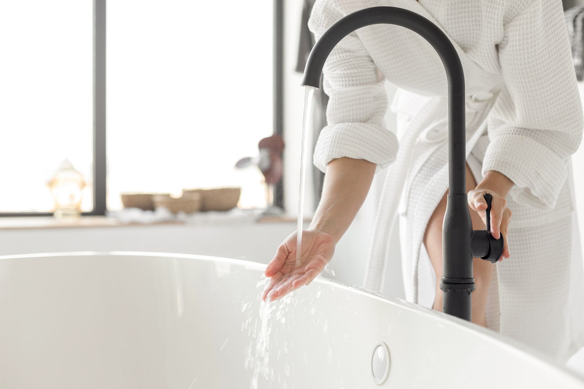 Person in white robe checks the water temperature from a black faucet over a white bathtub.