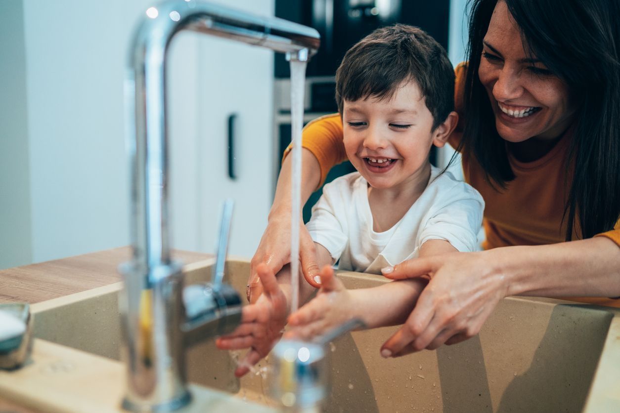 Woman and child washing hands together at a kitchen sink. Both smiling and laughing.