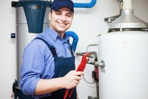 Smiling plumber in blue overalls and cap holding a wrench, near water heater.