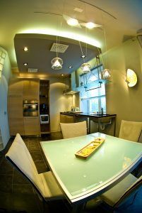 Modern kitchen with glass table, pendant lights, and light wood cabinets.