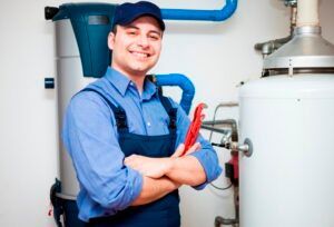 Smiling plumber in blue overalls, holding wrench, stands near water heaters.
