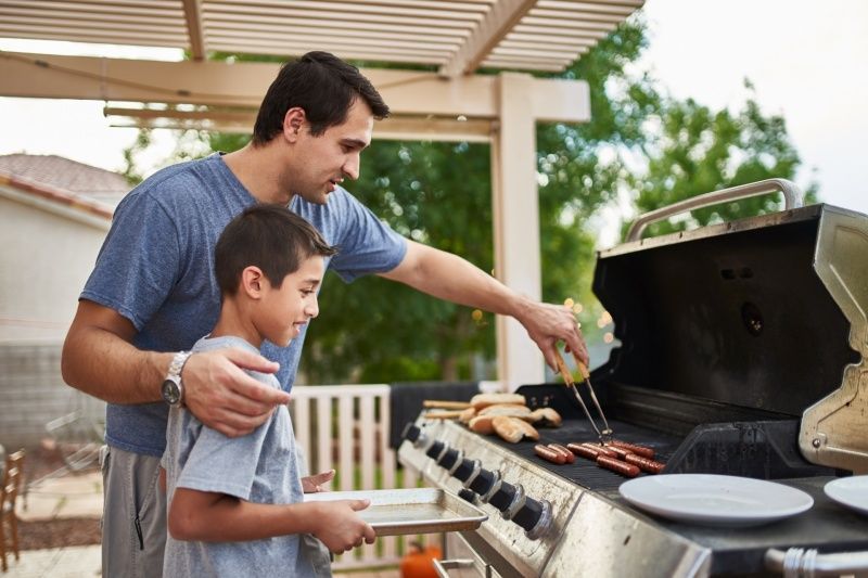 Father and son grilling hot dogs outdoors, enjoying a sunny day.