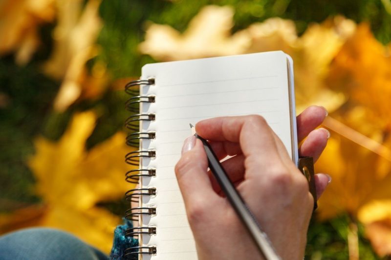 Hand writing in spiral notebook with pencil, fall leaves in background.