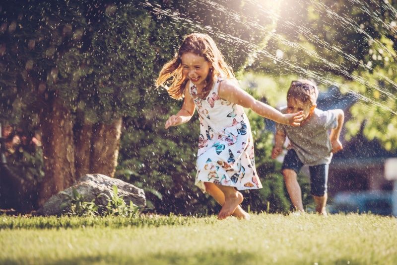 Children playing in a sprinkler on a sunny lawn, with a girl laughing and a boy running.