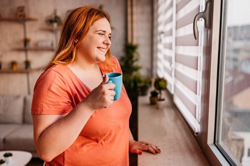 Woman with red hair smiles, holding a blue mug by a window with blinds.