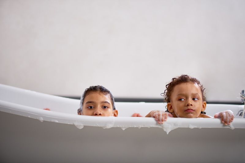 Two young children in a white bathtub, heads peeking over the edge, looking at the camera.