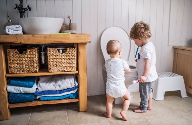 Two young children investigating a toilet in a bathroom; one is holding a toothbrush.