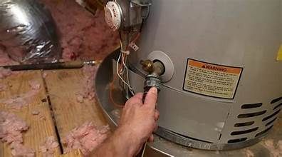 A person's hand is touching a water heater's drain valve in a utility room with pink insulation.