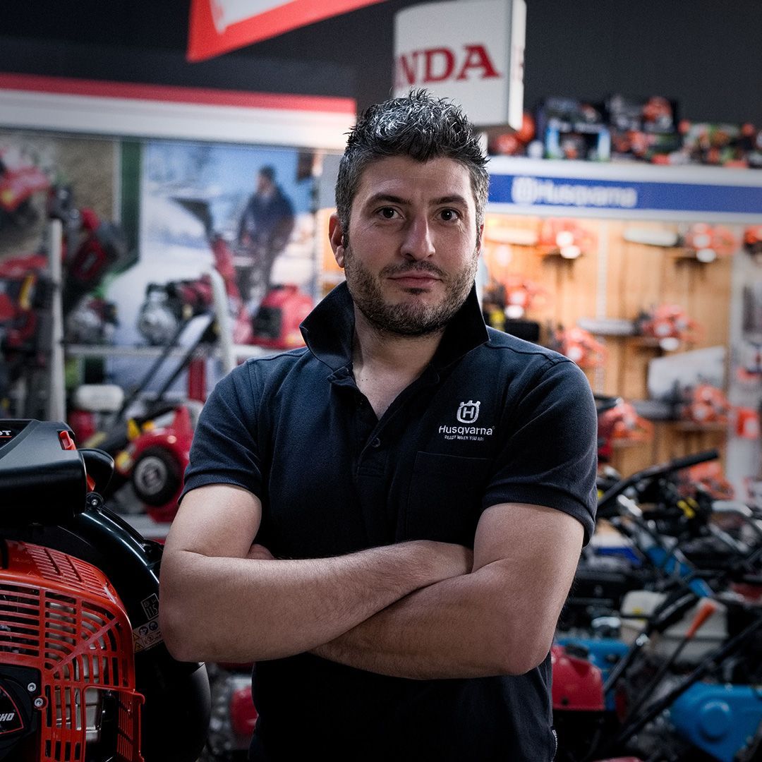 Uomo con le braccia incrociate, in un negozio di attrezzature da giardinaggio, che indossa una camicia nera.