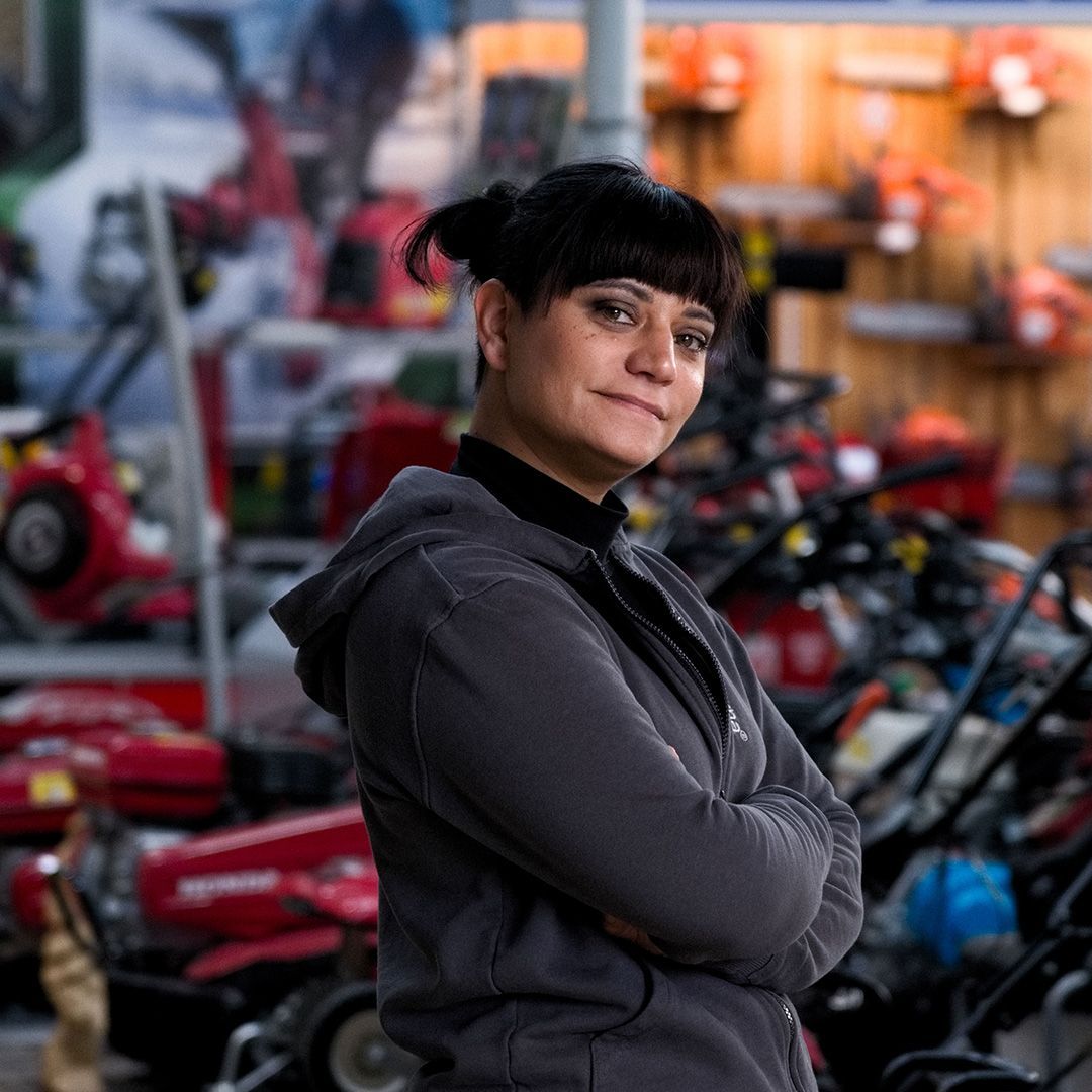 Donna con i capelli scuri, giacca grigia con cappuccio, braccia incrociate, sorridente in un negozio pieno di tosaerba.