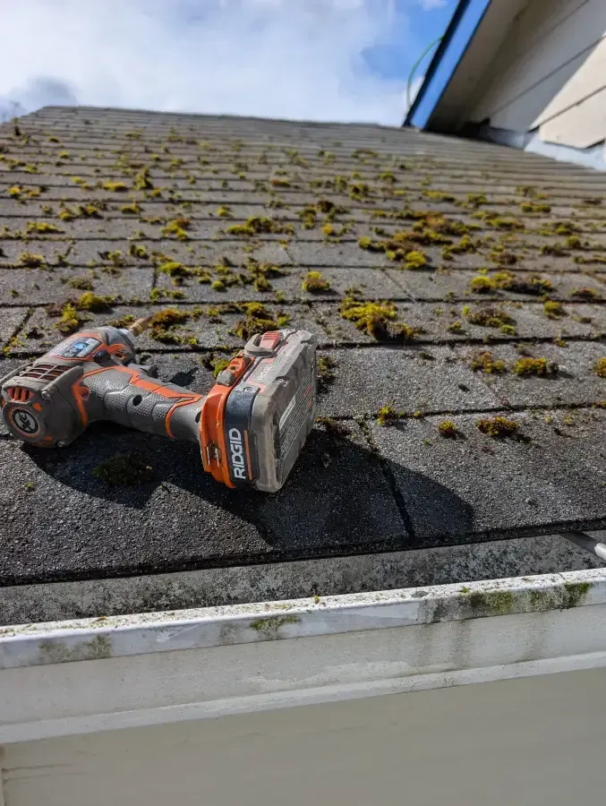 A power drill sits on a mossy, black shingled roof next to a white gutter.