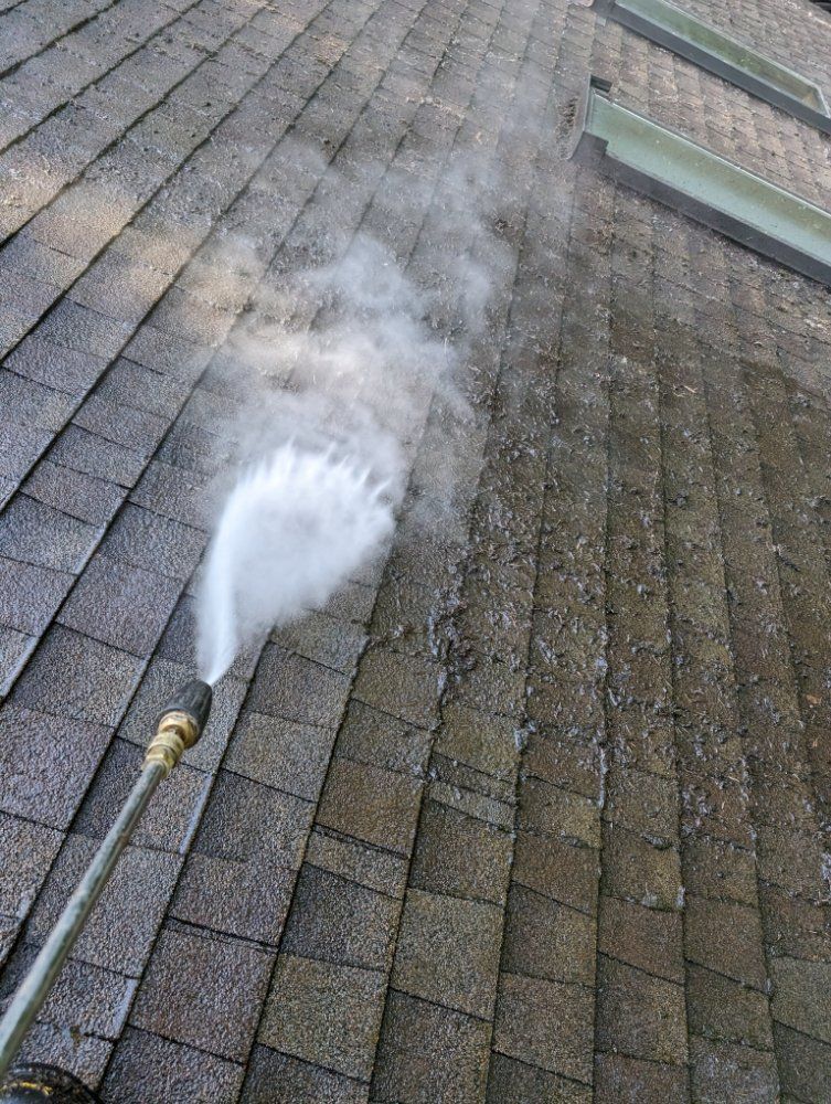 Pressure washing a weathered asphalt shingle roof, creating visible steam.