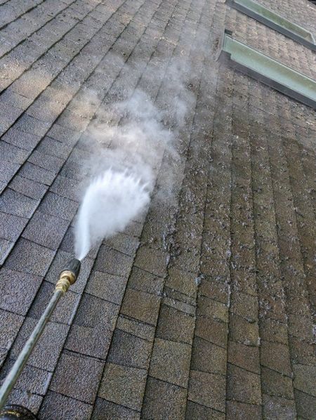 Pressure washing a weathered asphalt shingle roof, creating visible steam.