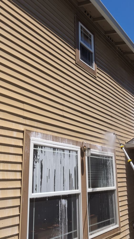 A person power washing a tan house, cleaning the windows, with white suds.