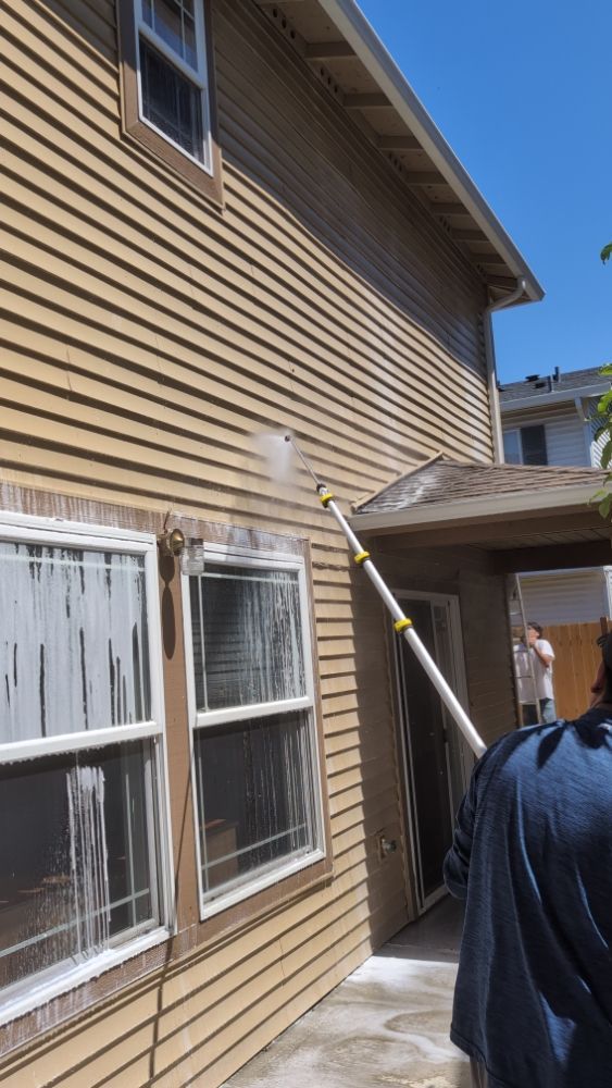 Person power washing the exterior of a beige house; soap suds cover the windows and siding.