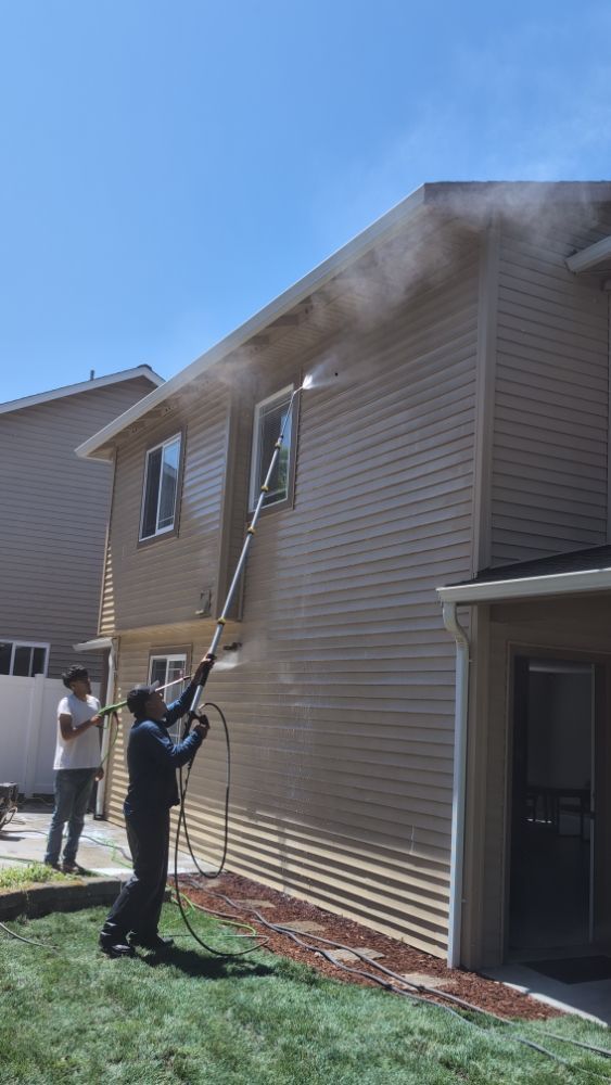 Two people power washing a beige two-story house on a sunny day. One sprays the siding.