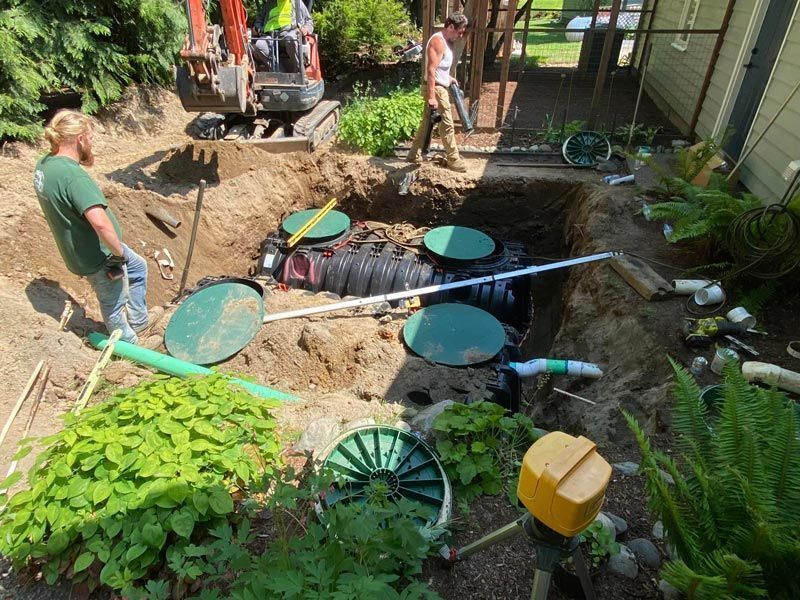 Construction workers installing a septic tank in a yard, using an excavator and hand tools.