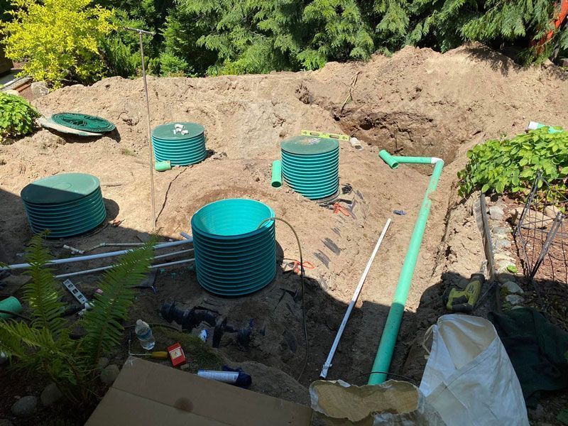 Installation of a septic system: green tanks and pipes in a dug-out area, surrounded by dirt and vegetation.